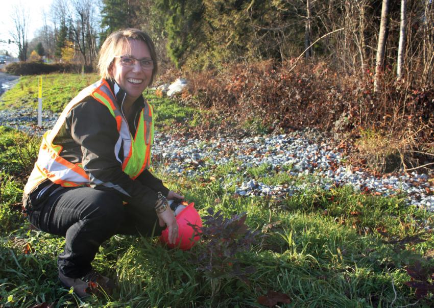Woman wearing safety vest crouches down in a grassy area, smiling at the camera