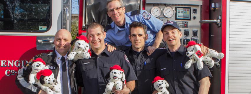5 firefighters hold stuffed dalmatians in front of a fire truck