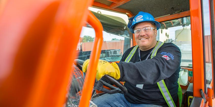 Man wearing safety gear sits behind the wheel of a forklift, smiling at the camera