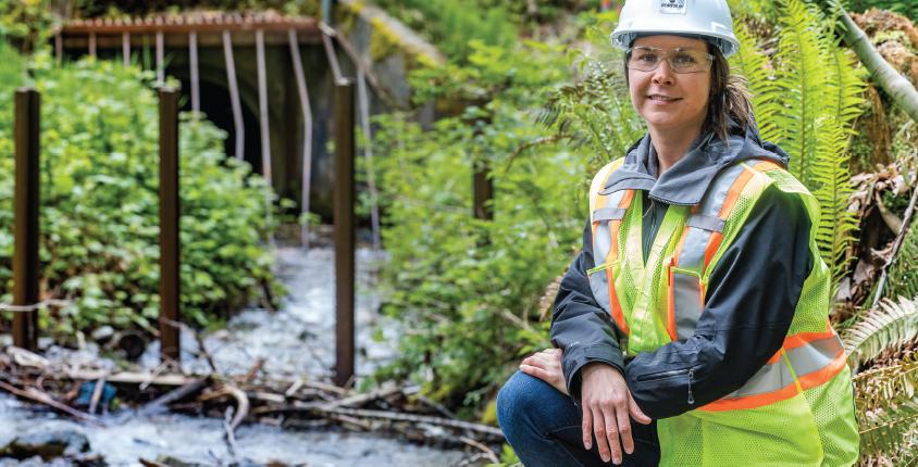 Woman kneels beside a creek wearing a hard hat and safety vest