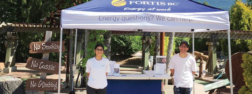 Two people stand under a FortisBC tent smiling at the camera