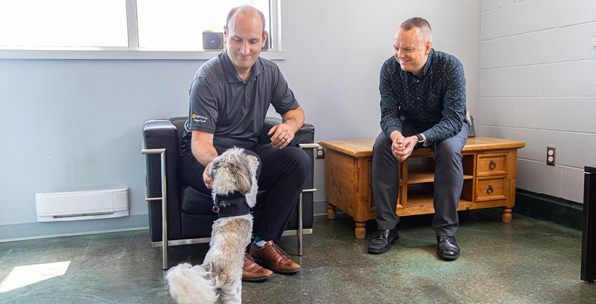 Two men sitting down, and a small dog jumping up on one man's knee