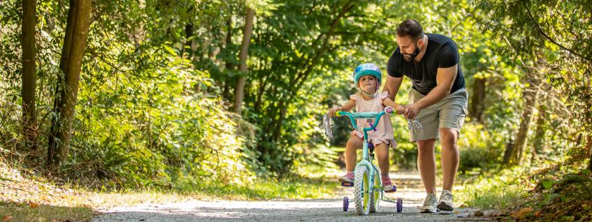Father helps daughter riding a bike with training wheels on the Riverview forest trail