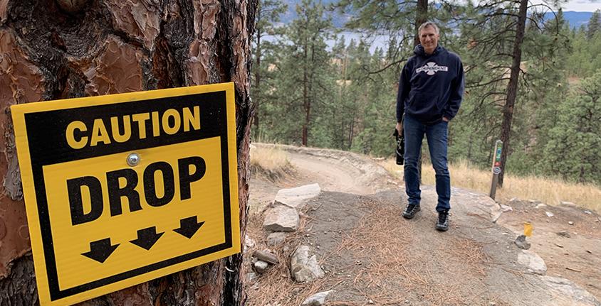 Man smiles at camera on bike trail beside a Caution: drop sign