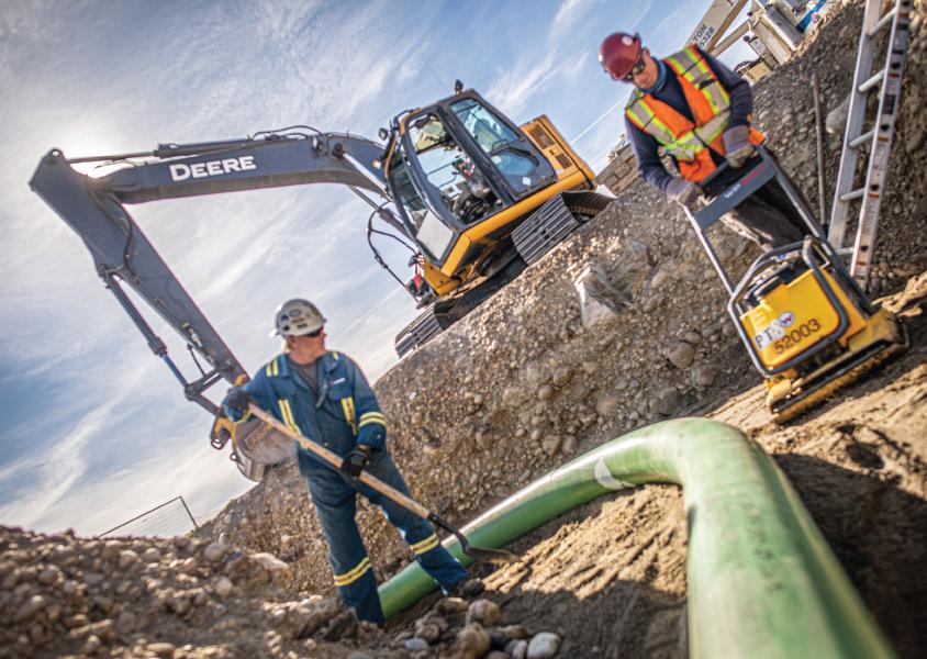 Two workers work in a pipeline trench, with an excavator in the background