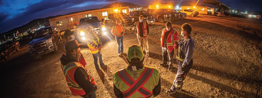 Workers stand in a circle, lit by headlights