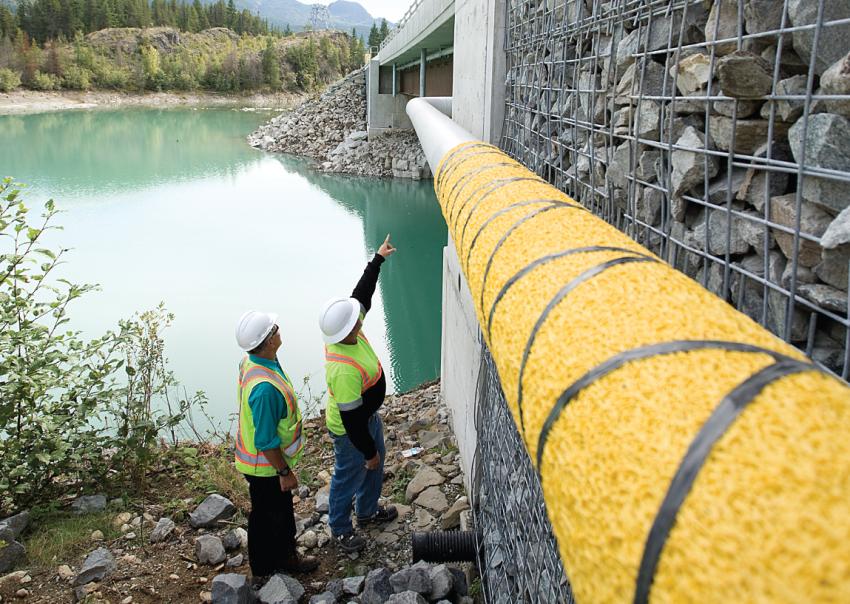 Two workers inspect a gas line beside a bridge