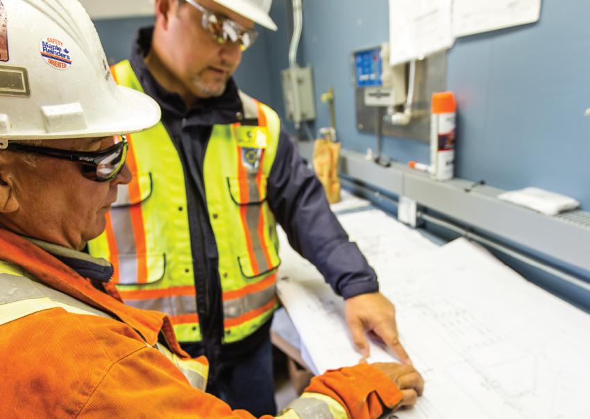 Two people wearing safety gear inspect blueprints