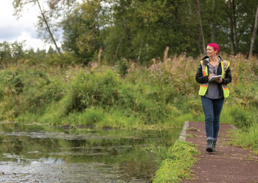 Caroline Astley walks beside a waterway holding a clipboard