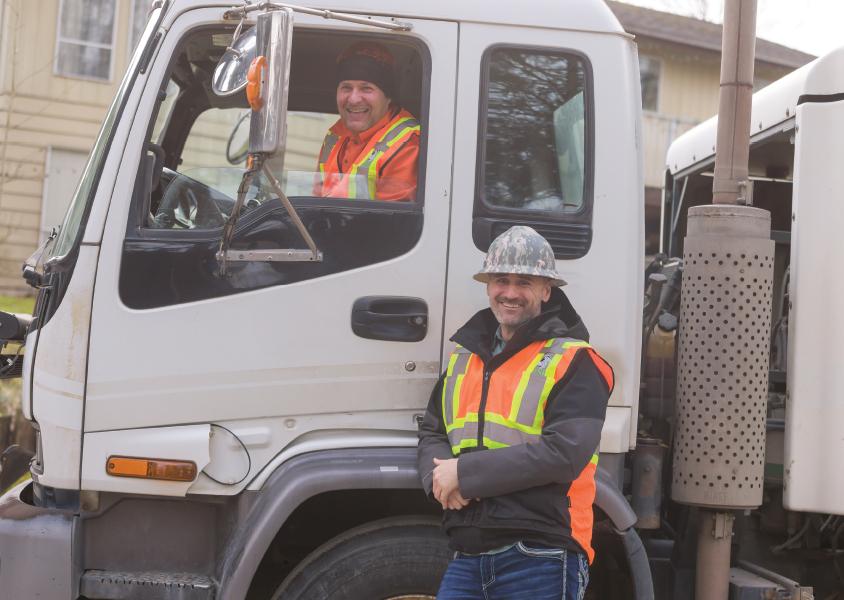 Man sits inside a truck and another stands beside the truck, both smiling at the camera
