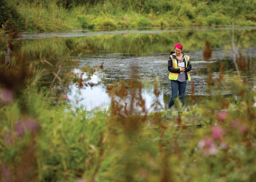 Woman wearing a safety vest stands at the edge of a lake looking down