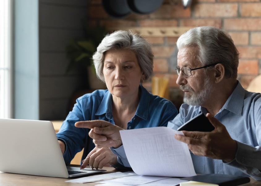 A senior couple looks at a computer screen and reviews paper documents at a table