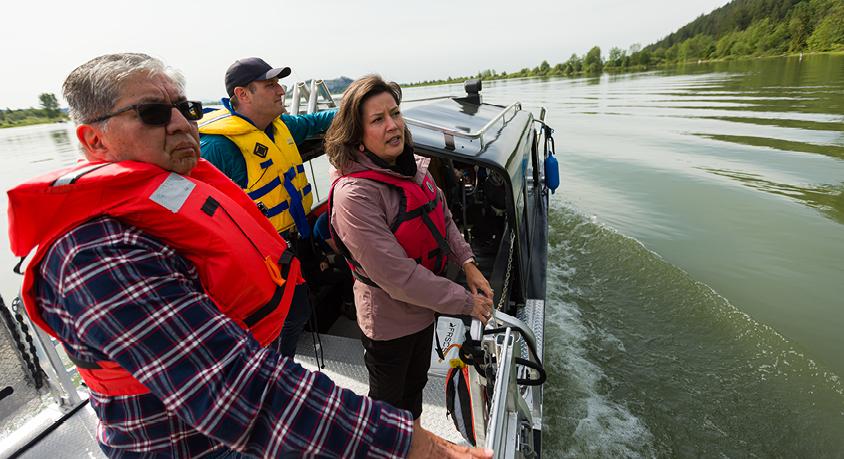 Three people wearing lifejackets stand on a boat in the Fraser River