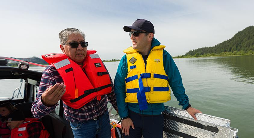 Two men wearing sunglasses and lifejackets talk on a boat