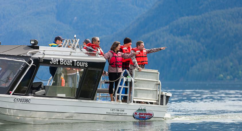 People are standing on a boat wearing lifejackets. One person is pointing, and the rest are looking in that direction. 