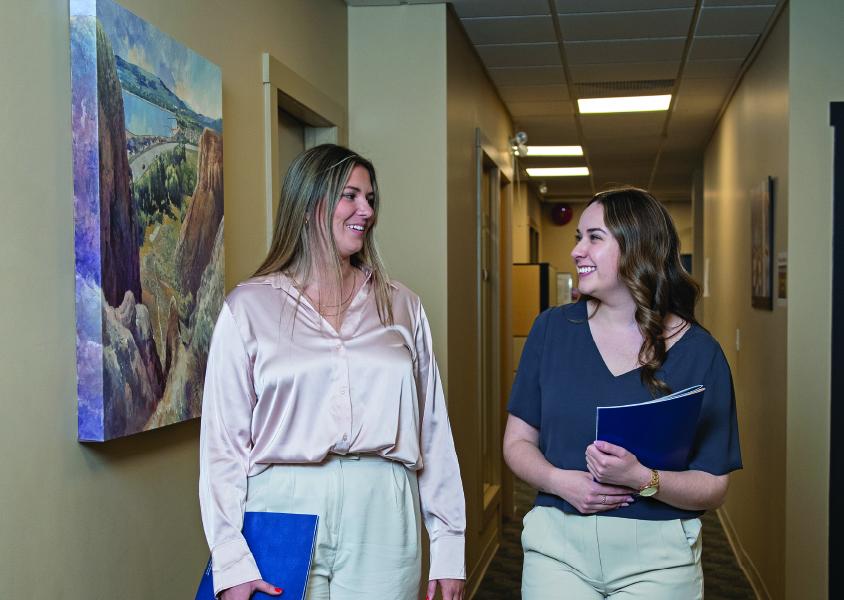 Two women walk down a hall looking at each other, talking