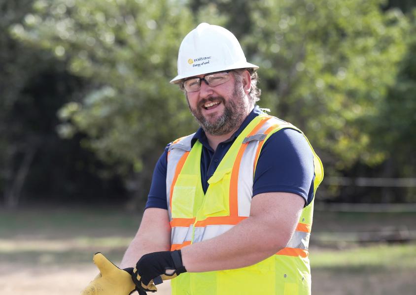 Matt Mason wearing a FortisBC hard hat, safety vest and work gloves smiles at something beyond the camera