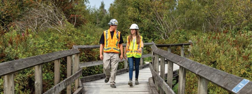 A man and a woman walk down a wooden boardwalk through a forested area. They are wearing safety vests and hard hats.