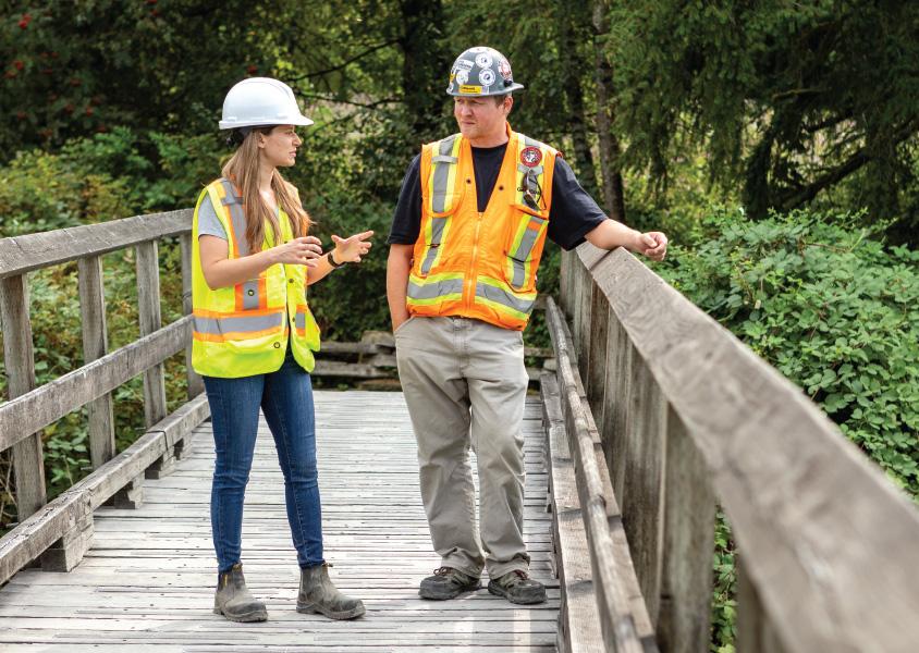 A man and a woman stand, talking on a wooden boardwalk in a forested area. They are wearing safety vests and hard hats.