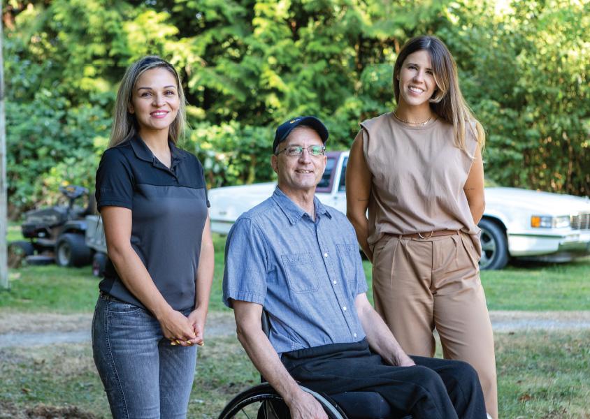 A man in a wheelchair sits between two women, all smiling at the camera