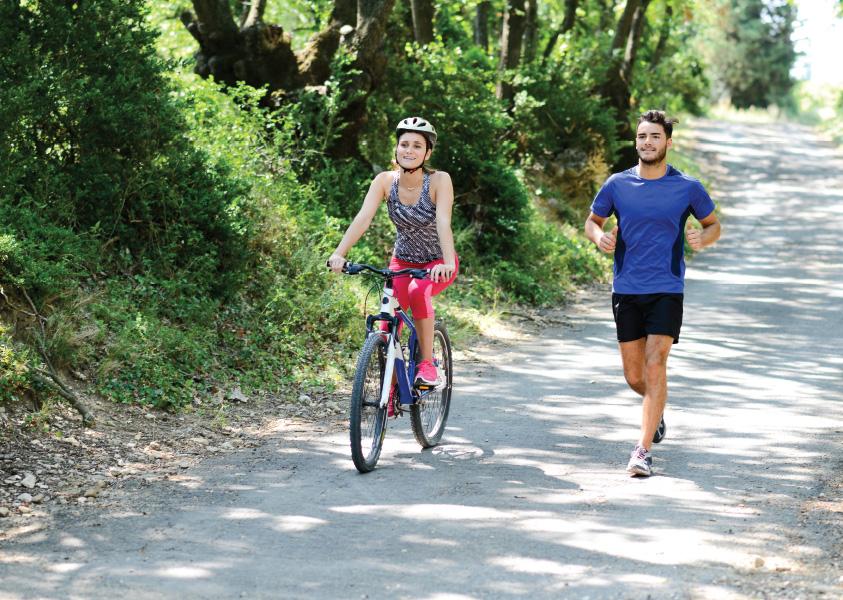 A woman on a bike and a man running down a sunny trail in the forest
