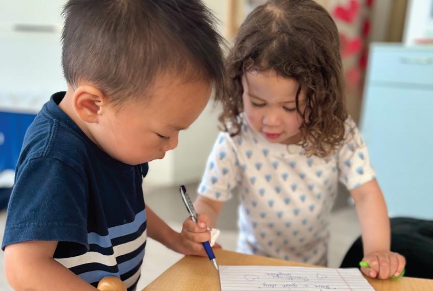 Two young children writing together at a table, focused on their task