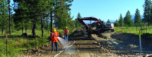 Two workers hose down a dirt roadway while an excavator works in the background