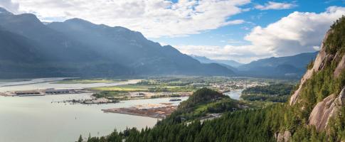 Aerial view of a coastal industrial town nestled between mountains, with a harbor, forested hills