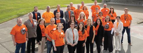 A group of approximately 25 people standing outdoors on a paved area. Most are wearing orange T-shirts with various designs and messages, while a few are in business attire. They are arranged in rows, smiling at the camera, with grass and steps visible in the background.